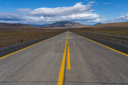 A long straight road with yellow lines leading towards mountains that crosses the Atacama desert in Chile. Sun with clouds in the blue sky.