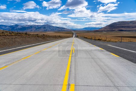 Foto de Un largo camino recto con líneas amarillas que conducen hacia montañas que atraviesan el desierto de Atacama en Chile. Sol con nubes en el cielo azul. - Imagen libre de derechos