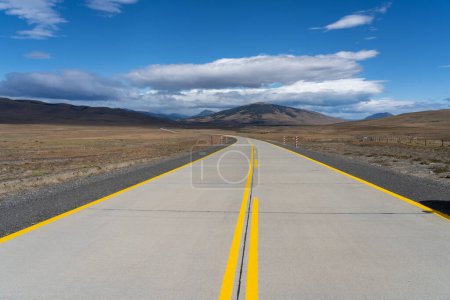 Foto de Un largo camino con líneas amarillas que conduce hacia montañas que atraviesan el desierto de Atacama en Chile. Sol con nubes en el cielo azul. - Imagen libre de derechos