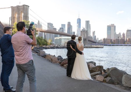 Photo for Wedding photo shoot in Brooklyn Bridge Park in NYC. Bride and Groom with Photographer and assistant. Sightseeing and picturesque place. - Royalty Free Image