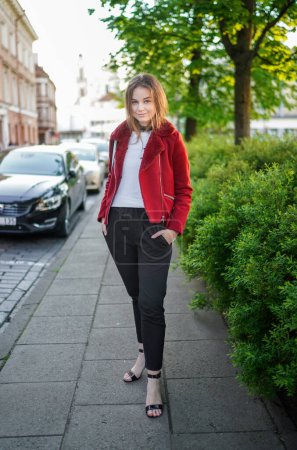 Foto de Beautiful Young Girl está de pie en el lado Paseo en el casco antiguo de Vilna, Lituania. Chaqueta roja y pantalones negros. Hermoso día de primavera. Sonriendo.. - Imagen libre de derechos
