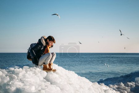 Téléchargez les photos : Young smiling woman sitting on a top of frozen sea ice blocks on a coast of the sea with a blue sky at the background, at frosty sunny day - en image libre de droit