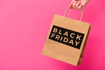 Black Friday, female hand holding brown craft shopping bag isolated on pink background. Black friday sale, discount, recycling, shopping and ecology concept