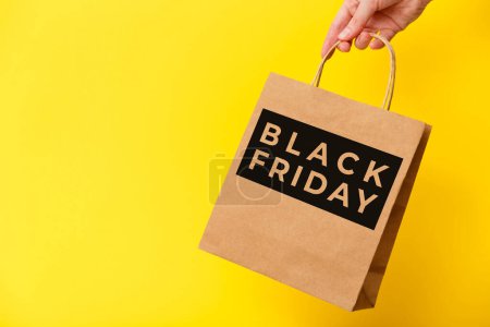 Black Friday, weibliche Hand mit brauner Bastel-Einkaufstasche auf gelbem Hintergrund. Black Friday Sale, Discount, Recycling, Shopping und Umweltkonzept