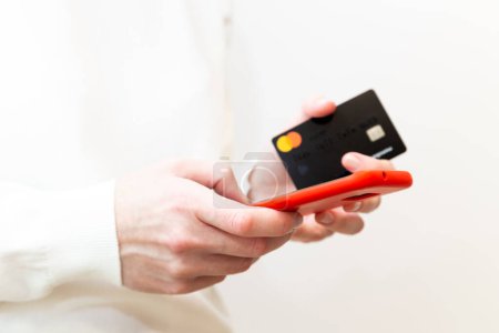 Téléchargez les photos : Kyiv, Ukraine - December, 2 2022: Close up of female hands holding Monobank Mastercard credit card and using smartphone in red case on white background. Paying securely online, using banking service. - en image libre de droit