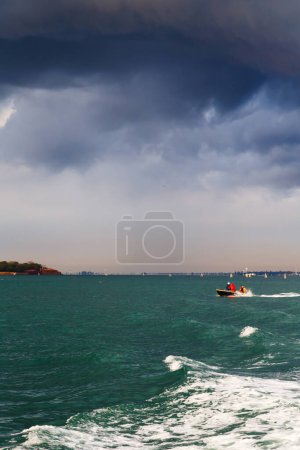 Mała łódź przepływająca obok wyspy La Grazia lub Santa Maria della Grazia w Weneckiej lagunie w burzliwej pogodzie z ciemnoniebieskim zachmurzonym niebem w tle.