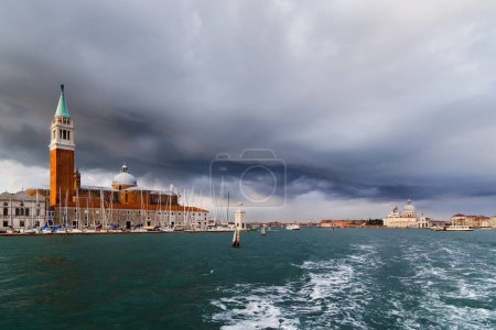 Venedig, San Giorgio Maggiore vom Wasser aus, vom Boot aus. Campanile Glockenturm, Kirche und Leuchtturm Faro der Insel San Giorgio Maggiore mit dunkelblauem, stürmischem Himmel im Hintergrund.