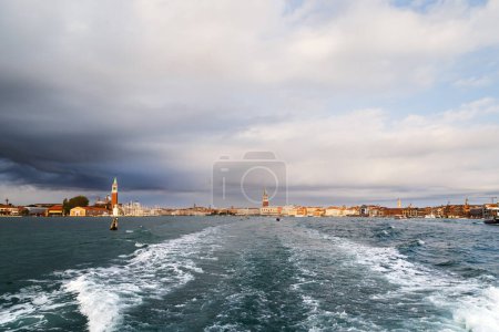 Venice panorama from sea and bright sea water trail behind small boat. San Giorgio Maggiore island, Saint Mark's Square, San Marco district view from water bus with dark blue stormy sky in background.
