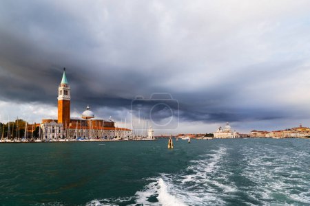 Campanile Bell Tower of San Giorgio Maggiore Island in Venice viewed from water bus. Church and lighthouse Faro of San Giorgio Maggiore island with dark blue stormy sky in background.
