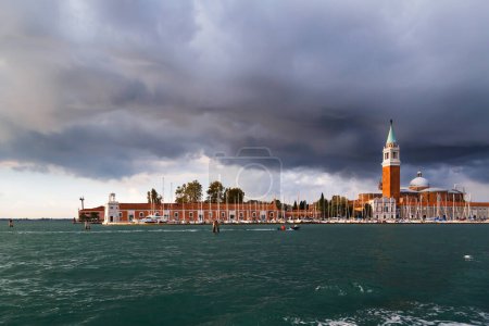 Wenecja, panorama wyspy San Giorgio Maggiore oglądane z wody, z łodzi. Campanile Bell Tower, kościół i latarnia morska Faro na wyspie San Giorgio Maggiore z błękitnym, burzliwym niebem w tle.