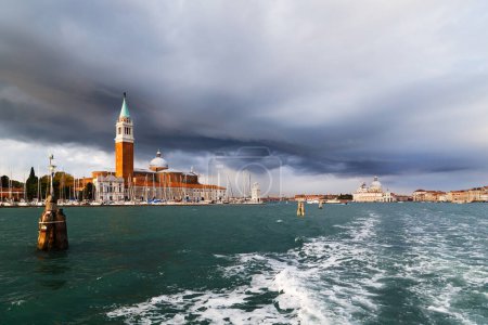 Venice panorama from the sea. San Giorgio Maggiore island view from water bus. Campanile Bell Tower, Church and lighthouse Faro of San Giorgio Maggiore island with dark blue stormy sky in background.