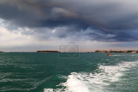 Blick auf Venedig, Italien von der venezianischen Lagune aus. Insel Giudecca und kleine Insel La Grazia oder Santa Maria della Grazia vom Wasserbus aus gesehen mit dunkelblauem stürmischem Himmel im Hintergrund.