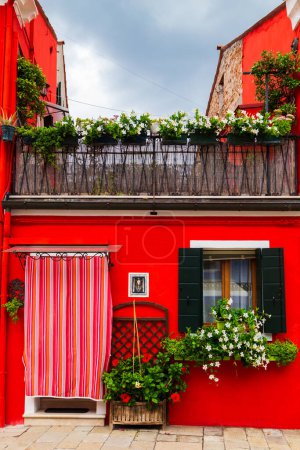 Helles traditionelles rotes Haus auf der Insel Burano, Venedig, Italien. Bunter Vorhang vor der Tür, hölzerne Fenster im alten Stil mit Rollläden und Mandevilla-Blumen auf der Fensterbank.