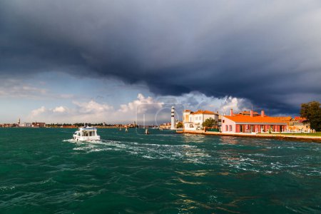 Murano Lighthouse or Faro di Murano is active lighthouse located on Murano island, Venice, Italy, in Venetian Lagoon on Adriatic Sea. View from water bus with dark blue stormy sky in background.