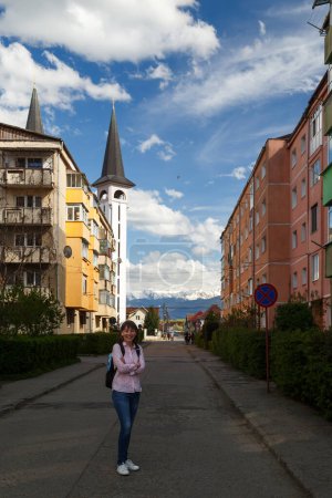 Young woman tourist at typical street in Romanian town of Avrig leading to Orthodox church with snowy Carpathian mountains in background, Romania. Soviet project of typical residential development.