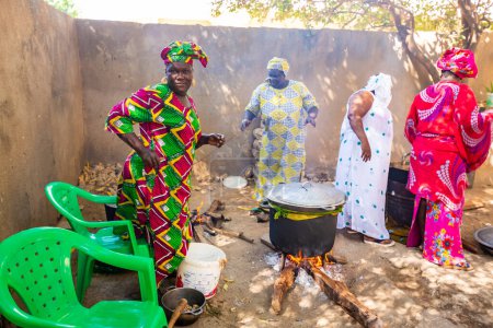 Photo for MBOUR, SENEGAL - Circa DECEMBER 2021. Unidentified women dressed with colorful traditional clothes cooking in outdoor kitchen with rudimentary utensil but great food. Poverty in Africa - Royalty Free Image