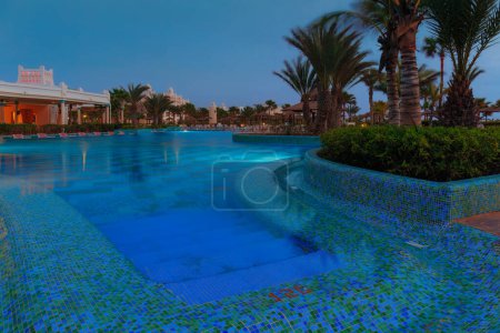 Foto de La elegante iluminación azul en la piscina del hotel crea un ambiente sereno, complementado con la silueta de palmeras y una vibrante escena del restaurante. Oasis nocturno. Foto de alta calidad - Imagen libre de derechos