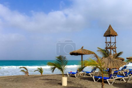 Beach day in Boa Vista : waves, a blue sky with white clouds, palm trees, sun loungers, a rattan beach umbrella and a wooden lifeguard tower.