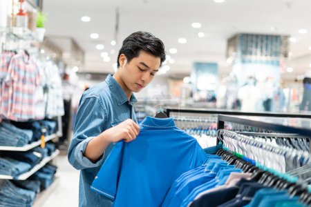 Foto de El hombre asiático inteligente está eligiendo la ropa en la tienda de ropa en el centro comercial, - Imagen libre de derechos