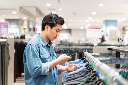 Foto de Asiático inteligente hombre usando su teléfono está recogiendo ropa en la tienda de ropa en el centro comercial, - Imagen libre de derechos