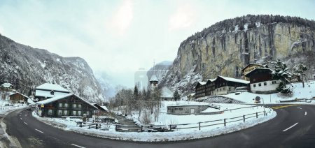 Foto de Panorama de invierno en un complejo turístico en los Alpes suizos. Un río fluye bajo un puente, y en los picos de la montaña cae una cascada, además de una antigua iglesia. - Imagen libre de derechos