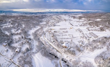 Drohnenblick über einem malerischen kleinen Dorf, das in einem Tal an einem kleinen Bach liegt. Wintersaison, jedes Haus, jedes Feld und jeder Baum ist mit Schnee bedeckt. Karpathien, Rumänien.