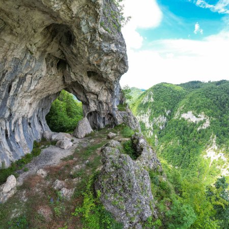 Viewpoint above mountains through a cave, eroded in a calcareous cliff on a rocky side. The tunnel has its stone walls covered with moss. The arches in the cave are uniting witha pillar. Carpathia