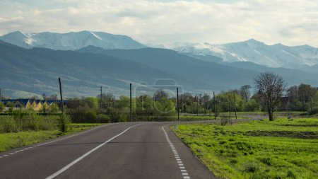 Eine asphaltierte Straße schlängelt sich durch eine idyllische Landschaft am Fuße verschneiter Berge. Am späten Nachmittag bringt die Sonne Wärme in die heitere Landschaft. Karpathien, Rumänien.