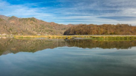 Ein ruhiger See mit Schilf und Sümpfen, die seine Ufer verengen. Der See liegt in einer ländlichen Gegend, umgeben von Hügeln und Buchenwäldern. Der Himmel spiegelt sich in seinem Wasser. Olt-Tal, Karpaten, Rumänien.
