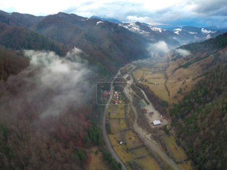 Drohnenpanorama über einem Tal in einer hügeligen Region. Wintersaison, die Berge sind verschneit. Der ländliche Raum ist von niedrigen Wolken bedeckt, die sich mit den Buchenwäldern vermischen. Karpathien,