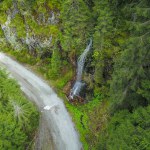 Drone view above a waterfall falling of a stone wall and flowing under a dirt road. The stream exits a spruce forest before falling into a cascade. Carpathia, Romania.