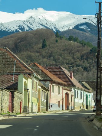 An asphalt street crossing a rural village in Eastern Europe. The houses are all built tight near each other, with huge gates. The countryside settlement is located at the feet of snowed mountains.