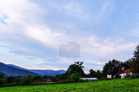 Photo for Farmhouses and a green meadow on the slope. The sun behind the clouds illuminates both the sky and the clouds with romantic light. The mountain range in the background - Royalty Free Image