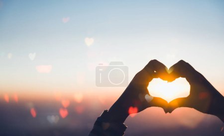 Female hands in the shape of a heart on an abstract tropical sunrise background with haert bokeh, sun light flares, waves, and blur. Copy space of joyful, freedom-loving travel concept.
