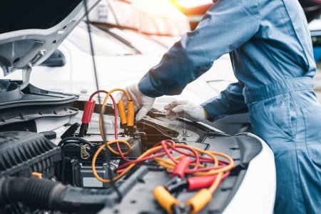 Photo for A professional mechanic in a blue uniform jump starts a car with jumper cables in a workshop setting - Royalty Free Image