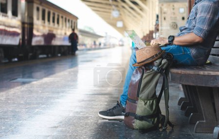 Norme numérique attend un train assis sur un banc à une gare, tapant sur un ordinateur portable et cherchant un emplacement sur une carte avec un sac à dos