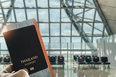 Großaufnahme der Hand eines Reisenden mit einem thailändischen Pass in einem hellen Flughafenterminal, im Hintergrund leere Sitze