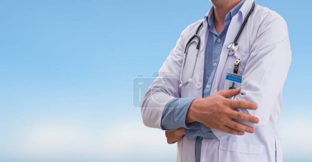 Medical doctor with stethoscope and ID badge standing confidently with arms crossed under a clear blue sky