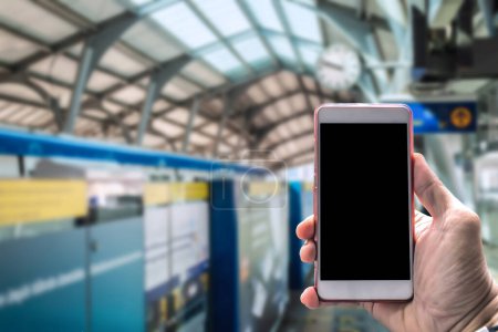 Close-up of a hand holding a blank-screen smartphone with a blurred train station background, suggesting travel and technology