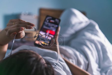 Ein Blick über die Schulter von einer Person im Bett, die ein Smartphone mit einer Brille in der Hand kontrolliert, was für eine moderne Morgenroutine steht