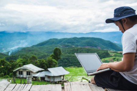 An individual uses a laptop outdoors, sitting on a wooden platform overlooking a lush mountainous landscape