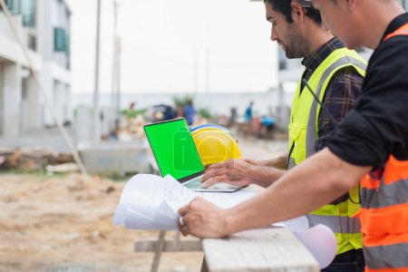 Two engineers with a laptop and blueprints at a construction site discussing project details