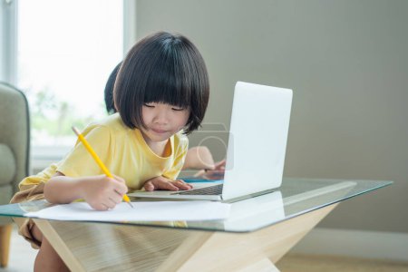 A young girl focuses intently on her studies using a laptop and writing in a notebook at a modern home setting