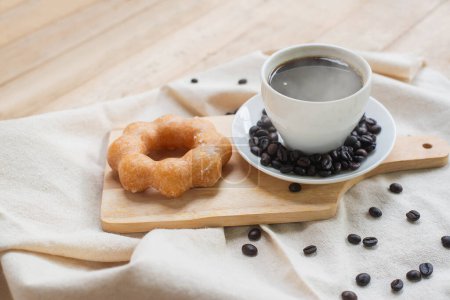 A freshly brewed cup of coffee paired with a glazed donut, coffee beans scattered on a rustic wooden table with linen