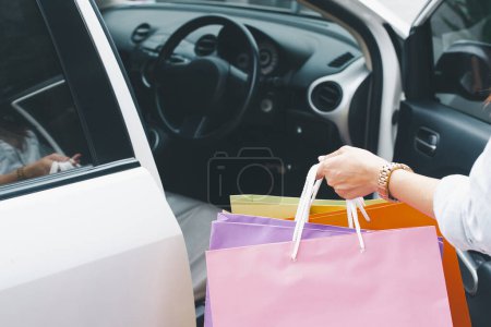 Un gros plan de la main d'une femme tenant des sacs colorés par une voiture, symbolisant un voyage shopping réussi