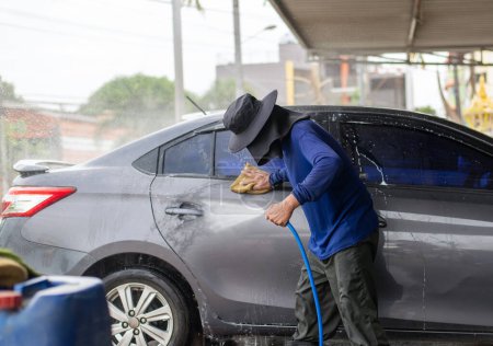 A worker in protective clothing washes a car at a car wash station, using a hose and sponge for cleaning.