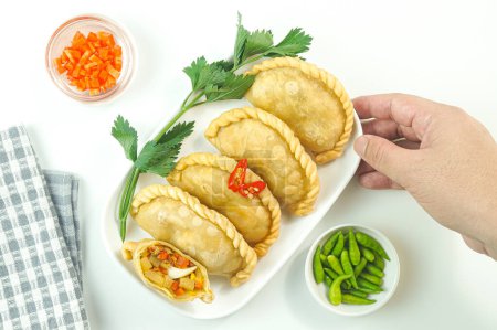 Téléchargez les photos : Pastels frits, en Indonésie connu sous le nom de Pastel goreng. Pâtisserie frite avec garniture de légumes, poulet et oeuf cuit servi avec du piment cru dans une assiette sur fond blanc à la main. Vue du dessus - en image libre de droit