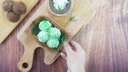 Téléchargez les photos : Kelepon gâteau ou klepon est un aliment traditionnel de l'Indonésie faite de farine de riz gluant noix de coco râpée sucre brun servi dans une assiette sur fond de bois sombre brillant à la main. Espace de copie de vue supérieure - en image libre de droit