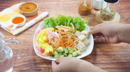 Téléchargez les photos : La salade de légumes crus ou Asinan Betawi est un aliment traditionnel indonésien composé de craquelins de sauce aux carottes et aux arachides au tofu et à la laitue au chou servis dans une assiette sur fond de bois sombre à la main. Gros plan - en image libre de droit
