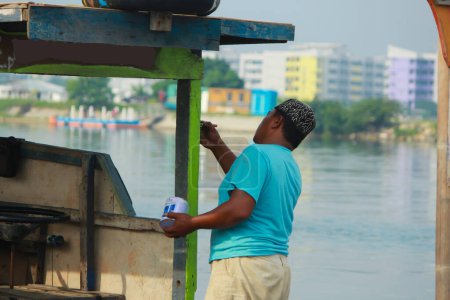 Téléchargez les photos : Un pêcheur peint son bateau sur les rives d'une rivière à Jakarta - en image libre de droit